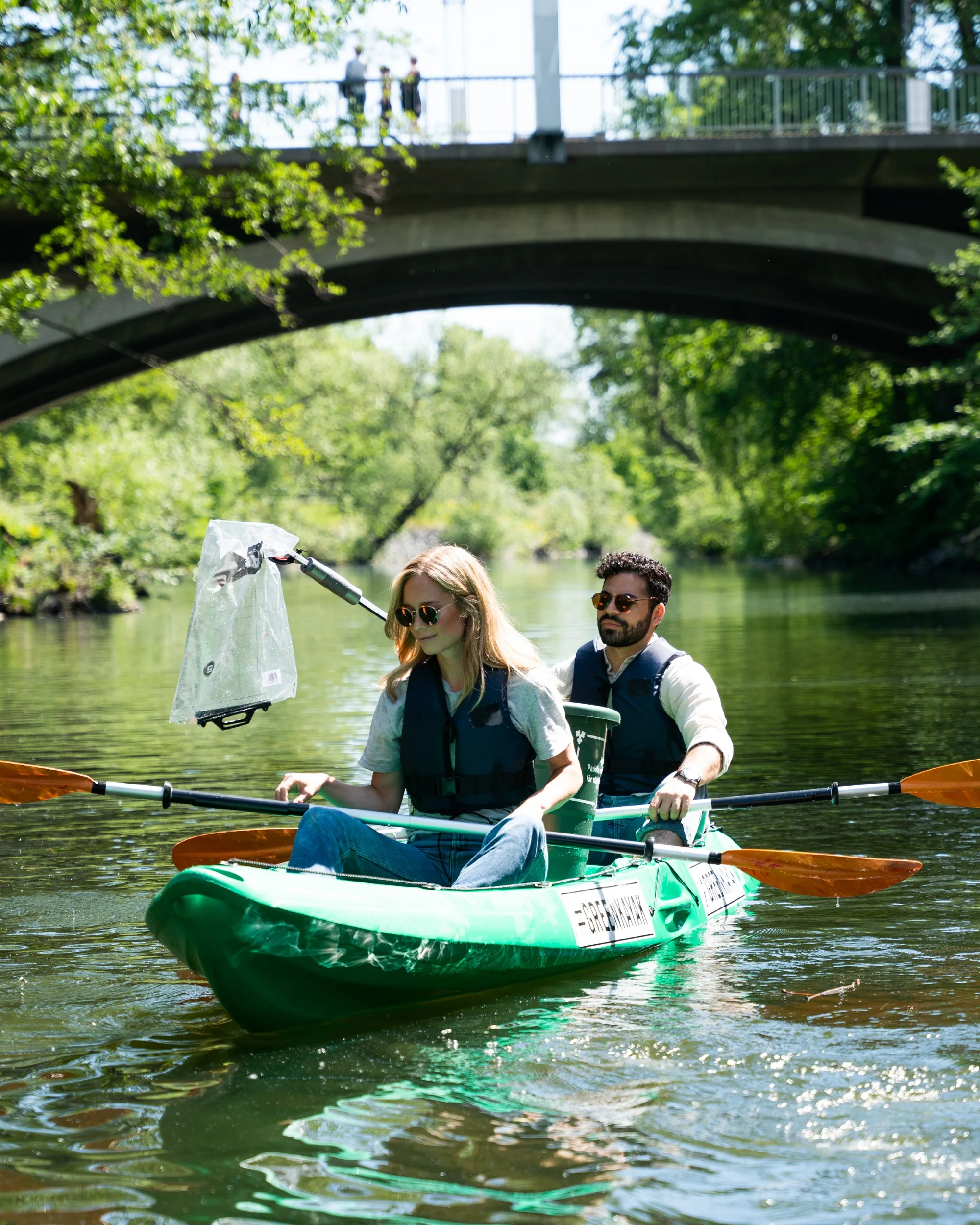 Two paddlers in a GreenKayak kayak under a bridge, holding a litter net